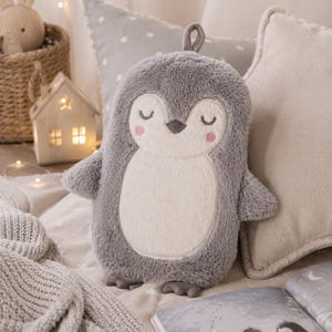 Puede incluir: Una funda de peluche para bolsa de agua caliente con forma de pingüino, hecha de piel sintética suave en color gris y crema. El pingüino tiene ojos cerrados bordados, un pequeño pico gris oscuro y dos mejillas rosadas. Incluye una pequeña trabilla.