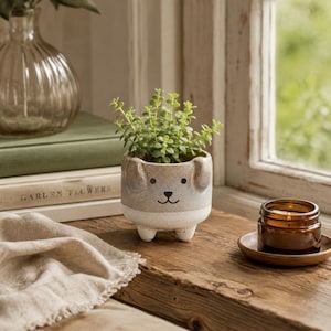 May include: A ceramic planter shaped like a dog with floppy ears, featuring a gradient of beige and white with a simple black painted face. The planter holds a small green succulent plant and stands on three short, rounded feet on a wooden surface.