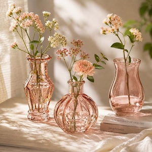 Könnte beinhalten: Drei hellrosa Glasvasen in verschiedenen Formen und Größen, die jeweils zarte Stiele mit kleinen rosa und weißen Blumen enthalten. Die Vasen haben strukturierte Oberflächen, darunter gerippte und facettierte Designs, und sind mit klarem Wasser gefüllt.