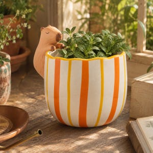 Peut inclure: Un pot de fleurs en céramique avec une figurine de capybara brun clair fixée sur le côté. Le pot a une forme arrondie avec un bord festonné et un motif de rayures verticales orange et jaunes sur fond blanc, contenant une plante verte.