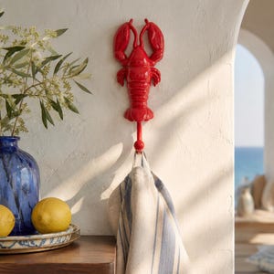 May include: A bright red wall-mounted hook shaped like a lobster, featuring a detailed crustacean design with two large claws at the top and a curved hook extending from the bottom, currently holding a beige and blue striped fabric towel.