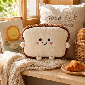 May include: A plush throw cushion shaped like a slice of bread with a dark brown crust and a cream-coloured centre. The cushion features an embroidered face with two black circular eyes, a small curved smile, and two pink circular cheeks.