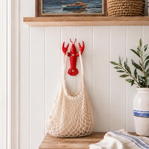 May include: A bright red lobster-shaped wall hook mounted on a white paneled wall. A beige cotton string mesh bag hangs from the lobster's claws. The hook features visible screw heads in the center and at the base for secure attachment to the wall.