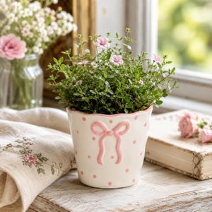 Könnte beinhalten: Ein weißer Keramik-Blumentopf mit einer erhabenen rosa Schleife auf der Vorderseite und einem Muster aus kleinen rosa Punkten. Der Topf hat einen gewellten oberen Rand und enthält eine grüne Pflanze mit kleinen rosa Blüten.