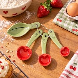 Può includere: Un set di quattro misurini in ceramica collegati da una catenella in metallo. I misurini presentano manici verde chiaro a forma di steli e foglie, con coppe rosse a forma di cuore. I misurini sono contrassegnati con 1 TBSP, 1 TSP, 1/2 TSP e 1/4 TSP.