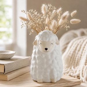 May include: A white ceramic sheep-shaped vase with a textured, wool-like surface and a smooth face featuring a black nose and mouth. The vase is filled with a bouquet of dried beige bunny tail grass and small white flowers, creating a soft display.