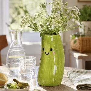 May include: A glossy, light green ceramic vase shaped like a pickle, featuring a simple black line-drawn smiling face with small pink cheeks and textured bumps on the surface, filled with a bouquet of white and green wildflowers.