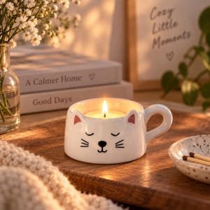 May include: A white ceramic mug shaped like a cat face with a lit candle inside. The mug features a painted black nose, mouth, and whiskers, with closed eyes and pink-tipped ears. A curved handle extends from the side of the round, glossy vessel.