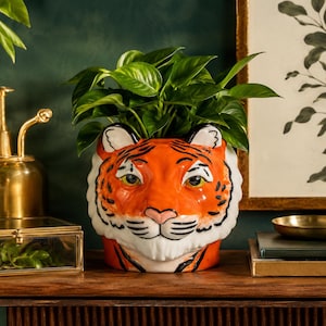 Puede incluir: Una maceta de cerámica con forma de tigre que presenta una cara de color naranja vibrante con rayas negras, mejillas blancas y una nariz rosa. La maceta contiene una planta de hojas verdes, con la cabeza del tigre formando el cuerpo principal.