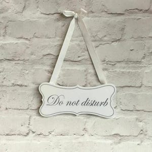 Può includere: Un cartello bianco decorativo con la scritta "Do not disturb" in elegante corsivo. Il cartello ha un bordo decorativo ed è sospeso da un nastro bianco legato a fiocco. Lo sfondo è un muro di mattoni bianchi.