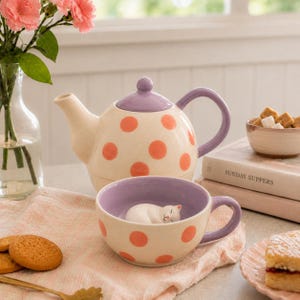 May include: A cream-coloured ceramic teapot and matching mug featuring orange polka dots and lavender accents. The teapot has a lavender lid and handle. The mug includes a small, sculpted white cat figurine resting at the bottom of the interior.