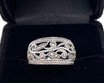 May include: A silver ring with an openwork design featuring a repeating pattern of swirling leaves and vines. The ring is displayed in a black velvet ring box against a white background. The ring has a detailed, textured appearance.