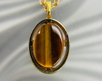 May include: A gold-colored necklace featuring an oval pendant with a tiger's eye stone. The stone is a warm brown color with a vertical stripe pattern. The pendant is set in a gold-tone frame and hangs from a delicate chain.