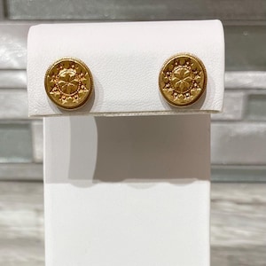 May include: A pair of gold-colored stud earrings. Each earring is round with a detailed floral design in the center, surrounded by a border of small star-like shapes. The earrings are displayed on a white stand.
