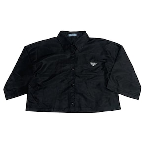 Puede incluir: Camisa negra abotonada con cuello y mangas largas. La camisa presenta un pequeño logotipo triangular blanco en el pecho. El logotipo de Prada es visible en el cuello. La camisa está hecha de un material brillante.