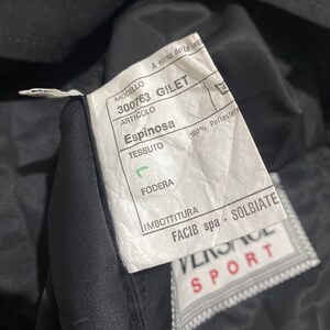 May include: A white tag with black text attached to a black garment. The tag reads "MODELLO 300763 GILET ARTICOLO Espinosa TESSUTO 100% Poliestere FODERA IMBOTTITURA FACIB spa - SOLBIATE" with a Versace Sport logo below.