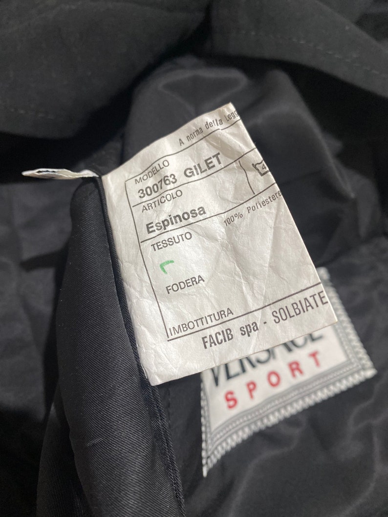 May include: A white tag with black text attached to a black garment. The tag reads "MODELLO 300763 GILET ARTICOLO Espinosa TESSUTO 100% Poliestere FODERA IMBOTTITURA FACIB spa - SOLBIATE" with a Versace Sport logo below.