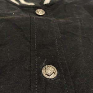 May include: Close-up of a black bomber jacket with a white striped collar and two silver buttons. The buttons feature a raised design of a head with a crown.