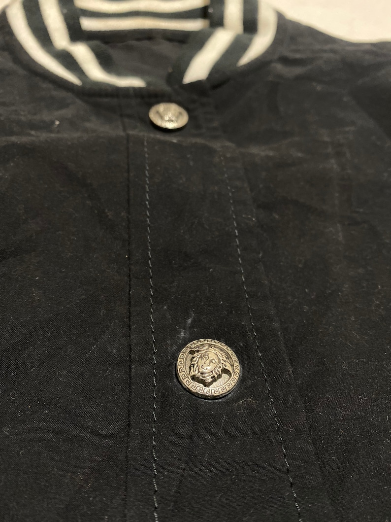 May include: Close-up of a black bomber jacket with a white striped collar and two silver buttons. The buttons feature a raised design of a head with a crown.