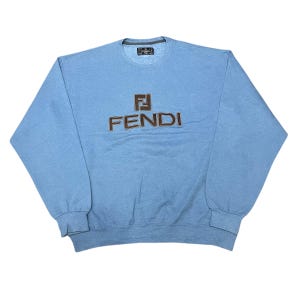 May include: A pale blue Fendi sweatshirt with a crew neck and long sleeves. The brown Fendi logo is embroidered on the front. The sweatshirt is made of a soft material.