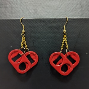 Puede incluir: Un par de pendientes rojos en forma de corazón hechos con materiales reciclados. Los pendientes están conectados por una cadena dorada.