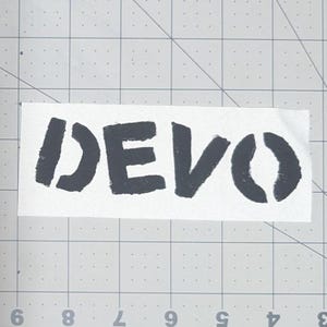 May include: A white rectangular sticker with the word "DEVO" printed in black, stencil-style lettering. The sticker is placed on a gray cutting mat with a grid pattern.