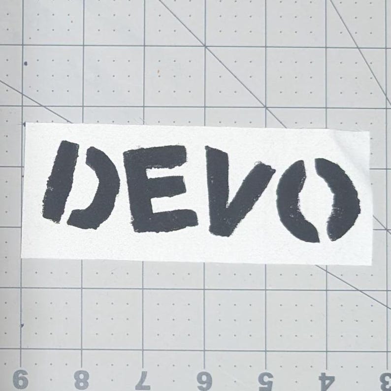 May include: A white rectangular sticker with the word "DEVO" printed in black, stencil-style lettering. The sticker is placed on a gray cutting mat with a grid pattern.