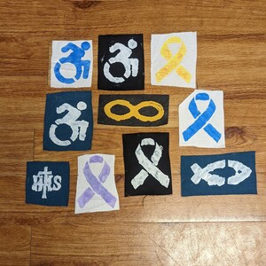 May include: Ten fabric patches with various designs, including infinity symbols, ribbons, and cross symbols. The patches are white, blue, yellow, and purple.
