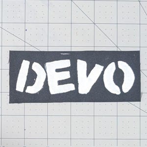 May include: A black rectangular patch with the word "DEVO" in white, stencil-style lettering. The patch is likely for clothing or accessories, and is set against a grid background.