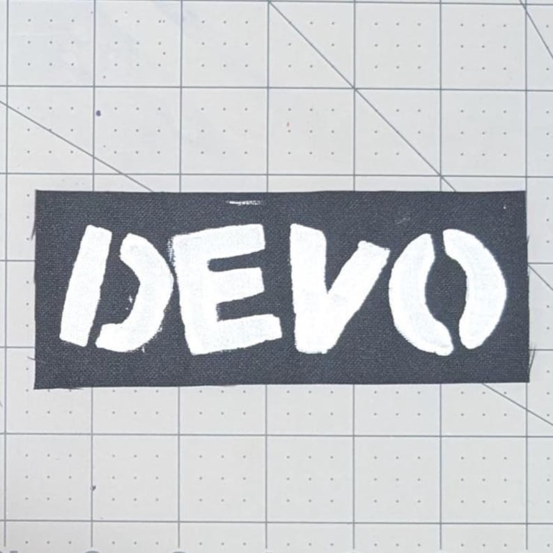 May include: A black rectangular patch with the word "DEVO" in white, stencil-style lettering. The patch is likely for clothing or accessories, and is set against a grid background.