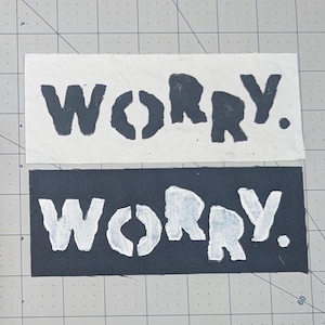 May include: Two fabric patches, one white and one black, each with the word "WORRY." stenciled in contrasting colors. The white patch has black lettering, and the black patch has white lettering. The word is in a bold, sans-serif font.