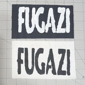 May include: Two rectangular patches, one black and one white, each displaying the word "FUGAZI" in bold, contrasting colors. The black patch has white lettering, while the white patch has black lettering. The patches are likely for clothing or accessories.