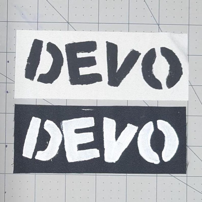 May include: Two fabric patches with the word "DEVO" printed in a stencil font. One patch is white with black lettering, and the other is black with white lettering. The patches are rectangular.