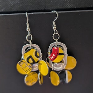 May include: A pair of earrings made from recycled aluminum beverage can tabs. The earrings are silver with yellow, black, and red accents. The tabs have text on them, including "OK + LL + 86", "OREGON", "Care", and "SM. A."