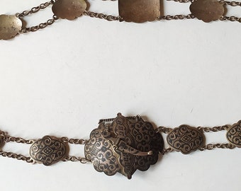Könnte beinhalten: Ein goldfarbener Kettengürtel mit einem dekorativen Verschluss und ovalen Gliedern. Die Glieder sind mit filigranen schwarzen Designs verziert.
