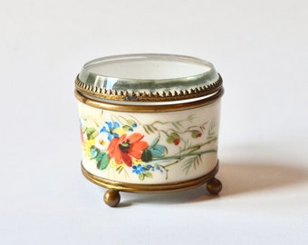 May include: A small, round, white porcelain trinket box with a glass lid. The box is decorated with hand-painted floral designs in shades of red, yellow, blue, and green. The box sits on three gold-colored feet.