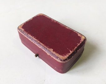 May include: A small, rectangular, burgundy-colored box with a gold hinge. The box is made of cardboard and has a worn, vintage look.