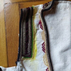May include: Close-up of the neckline of a white garment with intricate embroidery. The embroidery features a black, burgundy, silver, green, and yellow thread design.