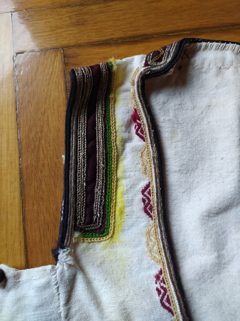 May include: Close-up of the neckline of a white garment with intricate embroidery. The embroidery features a black, burgundy, silver, green, and yellow thread design.