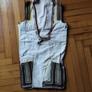 May include: A white sleeveless vest with intricate embroidery in shades of green, purple, and gold. The embroidery is around the neckline and the bottom hem.