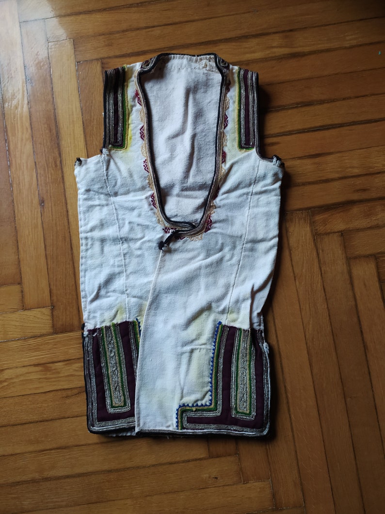 May include: A white sleeveless vest with intricate embroidery in shades of green, purple, and gold. The embroidery is around the neckline and the bottom hem.