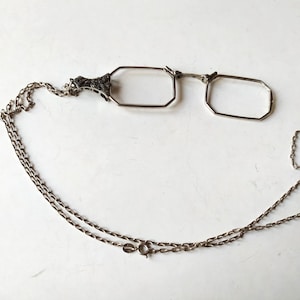 Op de afbeelding: Antieke zilverkleurige bril met achthoekige glazen en een decoratieve brug. De bril is bevestigd aan een delicate ketting, waarschijnlijk om om de nek te dragen. De ketting heeft een sluiting. De bril dateert waarschijnlijk uit het begin van de 20e eeuw.