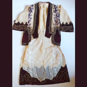 Può includere: Un costume tradizionale con un abito bianco ricamato e un gilet bordeaux con bordi dorati.