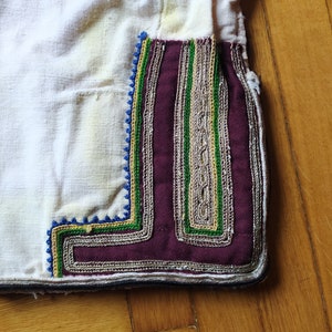May include: Close-up of a white fabric garment with a decorative trim. The trim is made of a burgundy fabric with silver, green, and blue embroidery.