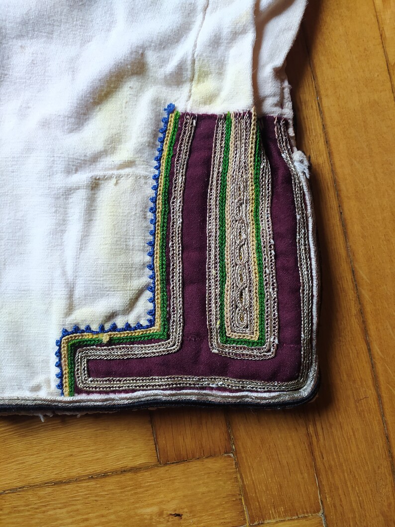 May include: Close-up of a white fabric garment with a decorative trim. The trim is made of a burgundy fabric with silver, green, and blue embroidery.