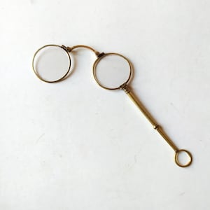 Könnte beinhalten: Antikes Monokel aus Messing mit Griff und kreisförmiger Schlaufe. Das Monokel hat zwei runde Gläser, die durch eine gebogene Brücke verbunden sind. Der Griff ist lang und schlank, mit einem kleinen Ring am Ende.
