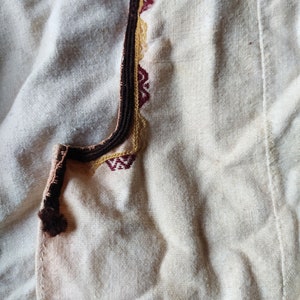 May include: A close-up of a white fabric with a brown, gold, and red embroidered trim. The fabric is textured and has a subtle sheen.