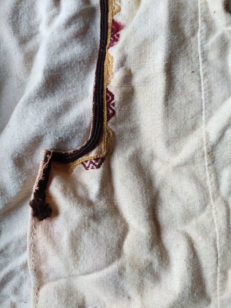 May include: A close-up of a white fabric with a brown, gold, and red embroidered trim. The fabric is textured and has a subtle sheen.