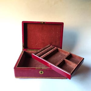 Könnte beinhalten: Eine rote Leder-Schmuckschatulle mit einem Scharnierdeckel und einem herausnehmbaren Fach mit Fächern. Die Schachtel hat ein goldenes Schloss und ein Schlüsselloch.