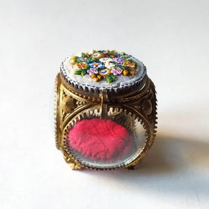 May include: An antique ring box with a floral mosaic lid. The box has a gold-coloured metal frame with clear glass sides. Inside, a red velvet cushion provides a place to hold rings. The mosaic features colourful flowers.